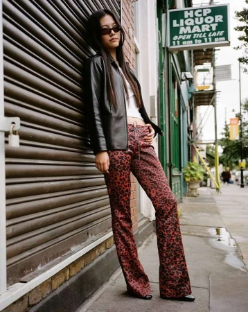Woman in sunglasses poses in front of roll-up door in a black jacket and red leopard print pants, standing on city sidewalk.
