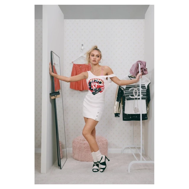 Young woman in a white dress posing in a dressing room with a mirror, clothing rack, and fluffy stool, creating a fashion forward scene.