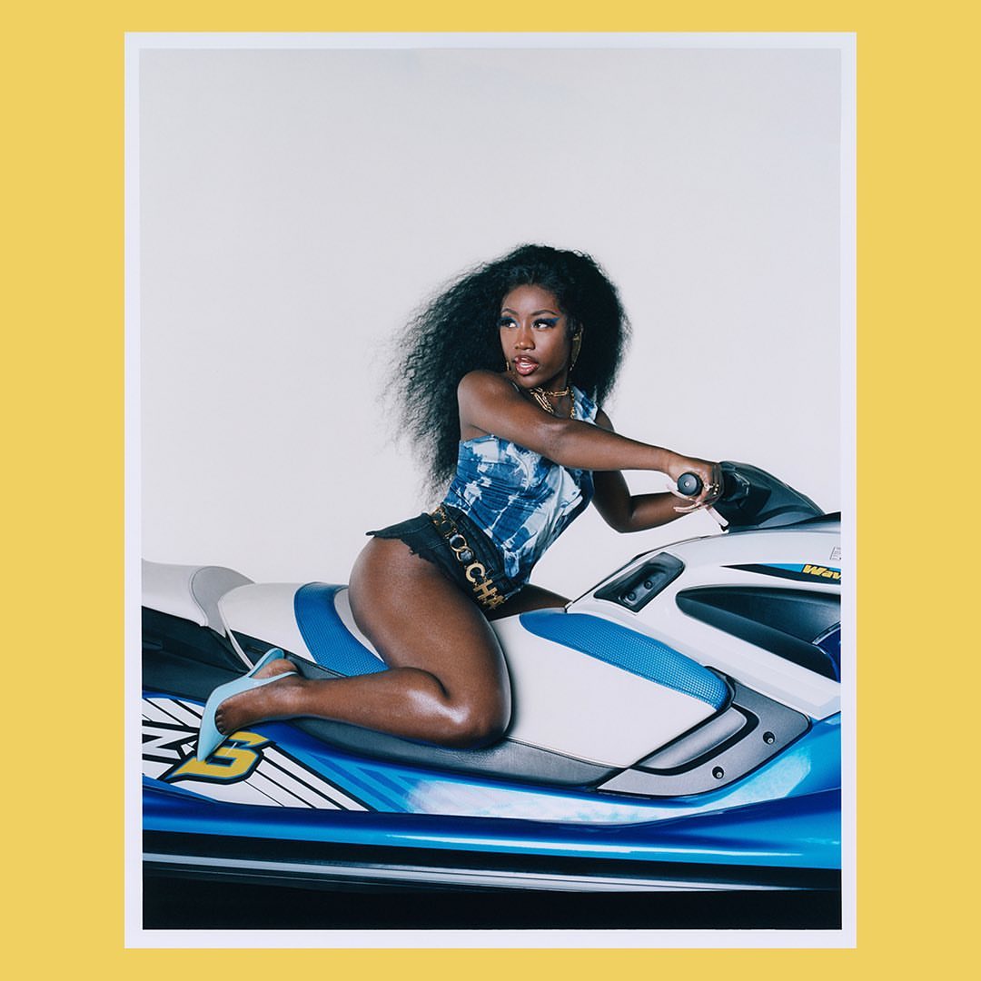 A woman with curly black hair and wearing a blue tie-dye top is sitting on a blue and white jet ski in a studio setting.