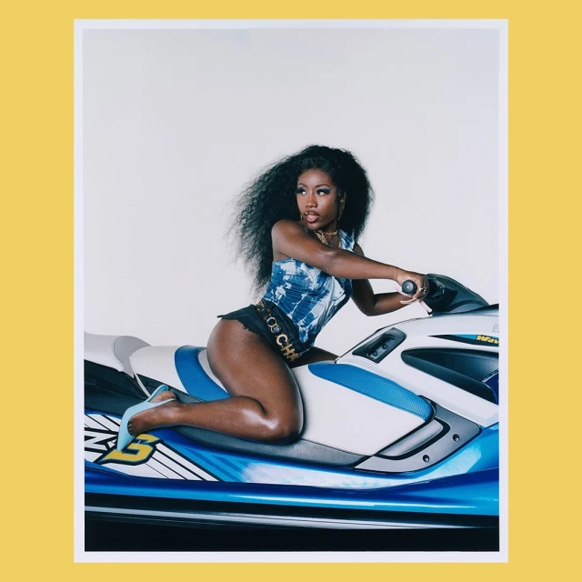 A woman with curly black hair and wearing a blue tie-dye top is sitting on a blue and white jet ski in a studio setting.