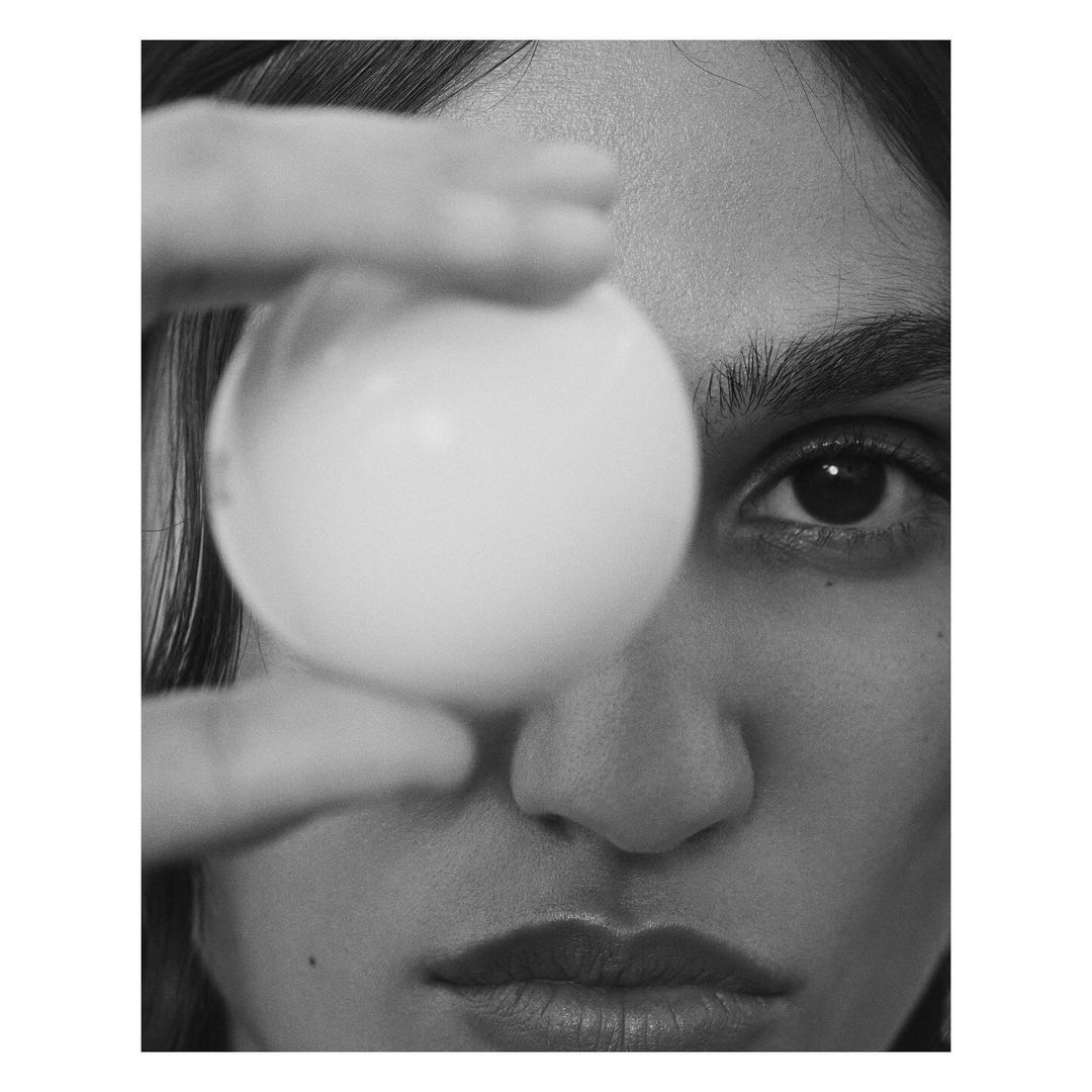 Monochromatic portrait of a woman holding a white sphere in front of her eye, creating an abstract and minimalist composition.
