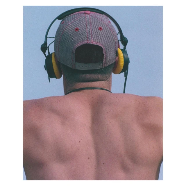 Back view of shirtless adult wearing a baseball cap and black headphones with yellow earpads against light blue sky backdrop.