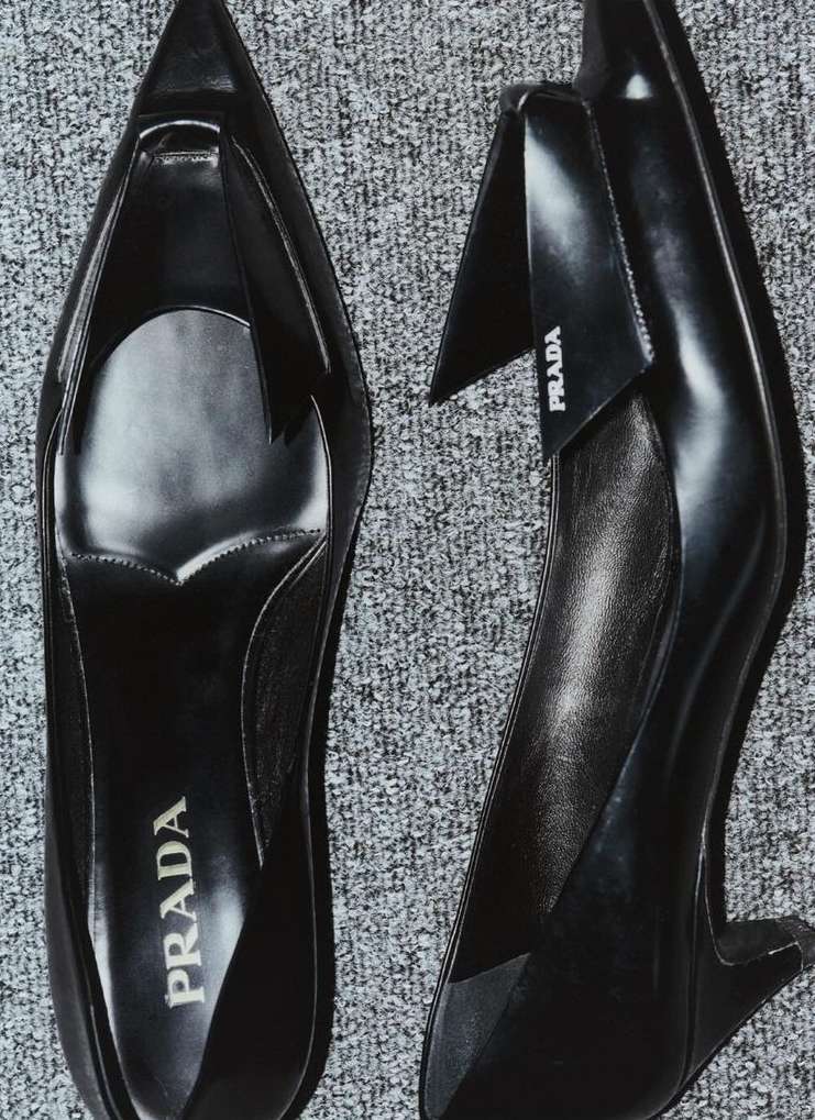 A pair of sleek black Prada heels is captured from a high angle against a textured gray background, highlighting their elegant design.