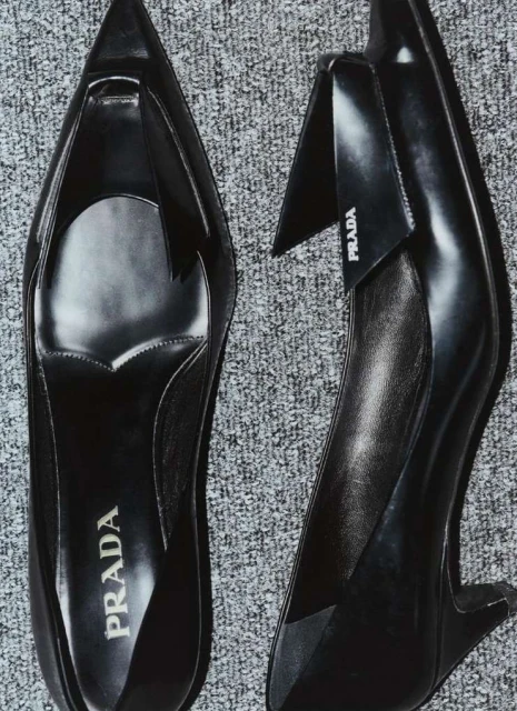 A pair of sleek black Prada heels is captured from a high angle against a textured gray background, highlighting their elegant design.