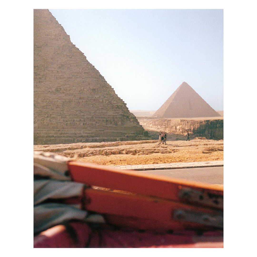 The Great Pyramid of Giza towers over a desert landscape. Three people walk in the distance, with part of a car visible in the foreground.
