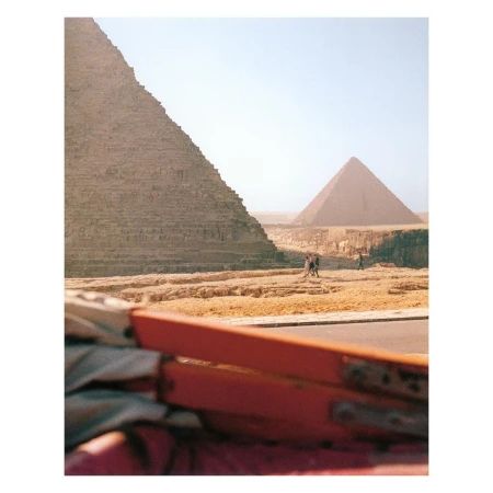 The Great Pyramid of Giza towers over a desert landscape. Three people walk in the distance, with part of a car visible in the foreground.