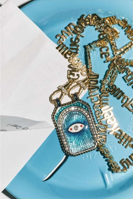 Close-up studio shot of a gold chain necklace with letter charms and a blue eye pendant on a light blue plastic surface with white paper.