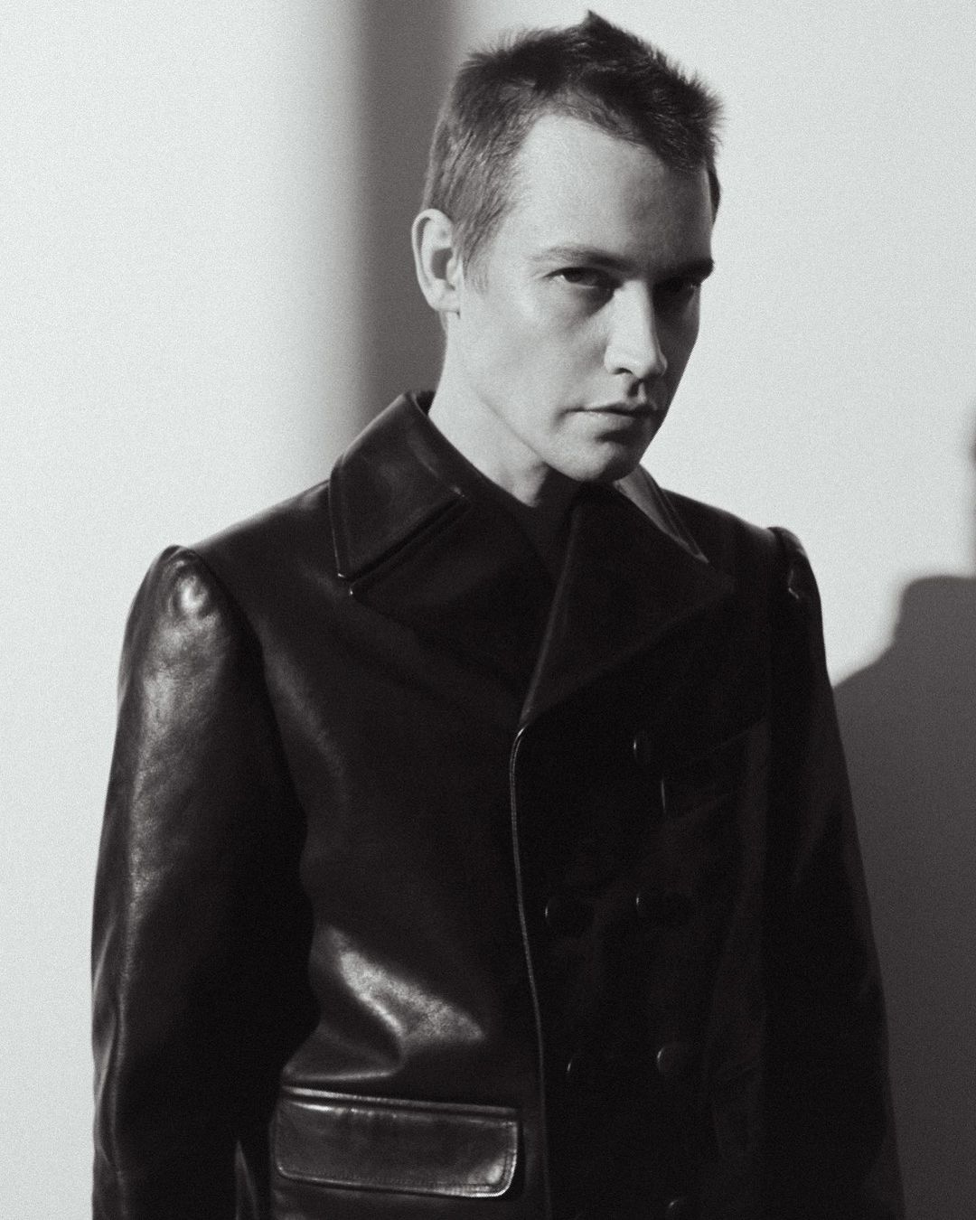 Black and white portrait of an adult male wearing a dark, double-breasted leather coat, looking at the camera with a neutral expression.