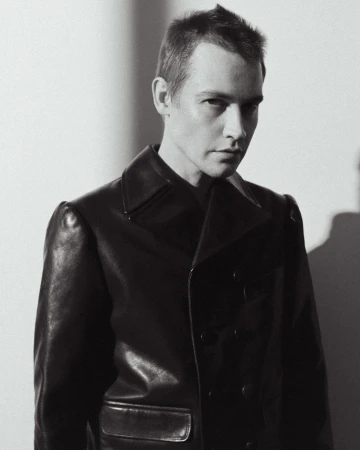 Black and white portrait of an adult male wearing a dark, double-breasted leather coat, looking at the camera with a neutral expression.