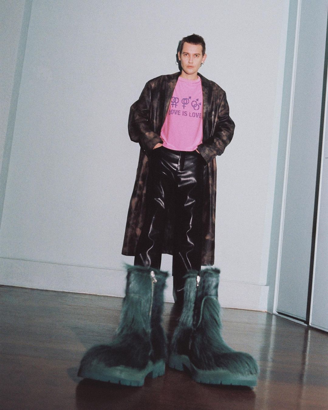 A full shot of a person wearing a pink t-shirt, long brown coat, black pants, and oversized furry blue-green boots in a studio.