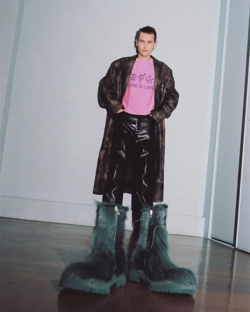 A full shot of a person wearing a pink t-shirt, long brown coat, black pants, and oversized furry blue-green boots in a studio.