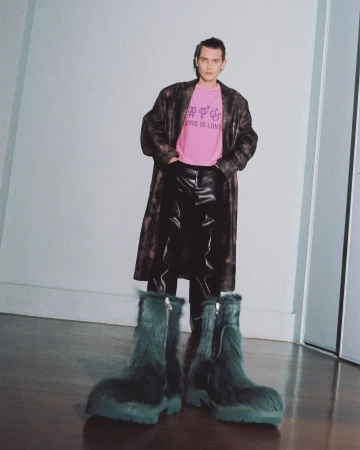 A full shot of a person wearing a pink t-shirt, long brown coat, black pants, and oversized furry blue-green boots in a studio.