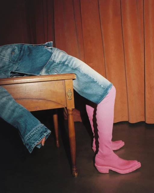 Person's lower half with pink boots leans against a wooden table, wearing a blue denim jacket, set against an orange curtain backdrop.