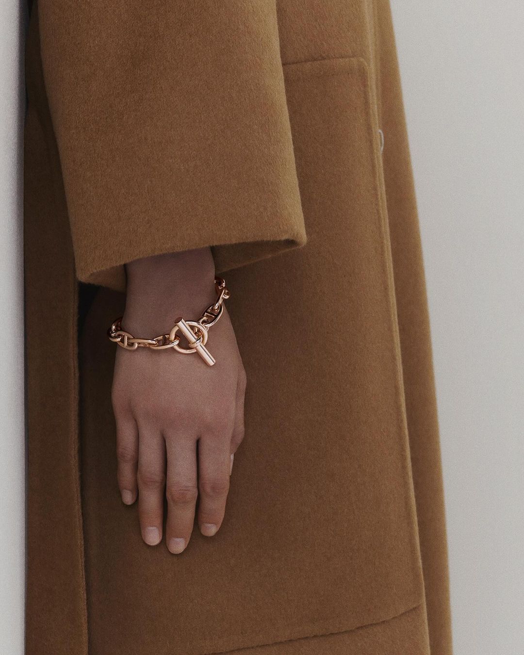 Close-up of a woman's hand wearing a gold chain bracelet against a tan wool coat and neutral background. Elegant jewelry fashion.