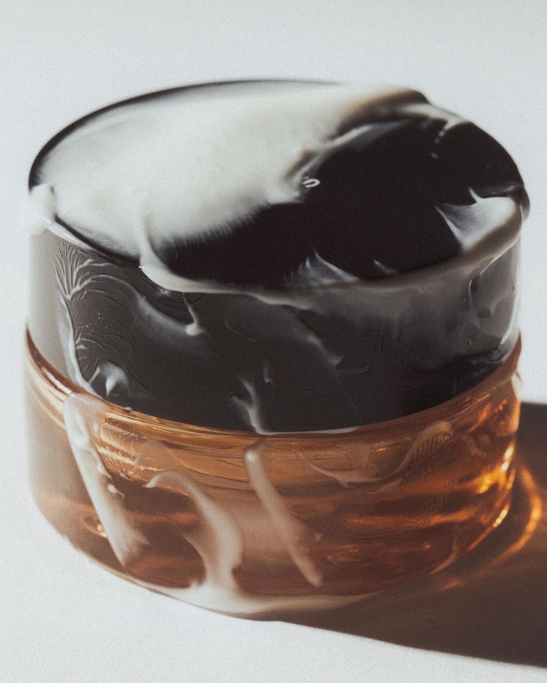 Close-up shot of a brown cosmetic jar with a black and white marbled lid, sitting on a white surface with soft lighting.