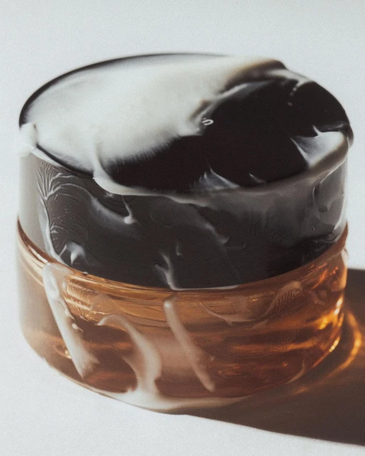 Close-up shot of a brown cosmetic jar with a black and white marbled lid, sitting on a white surface with soft lighting.