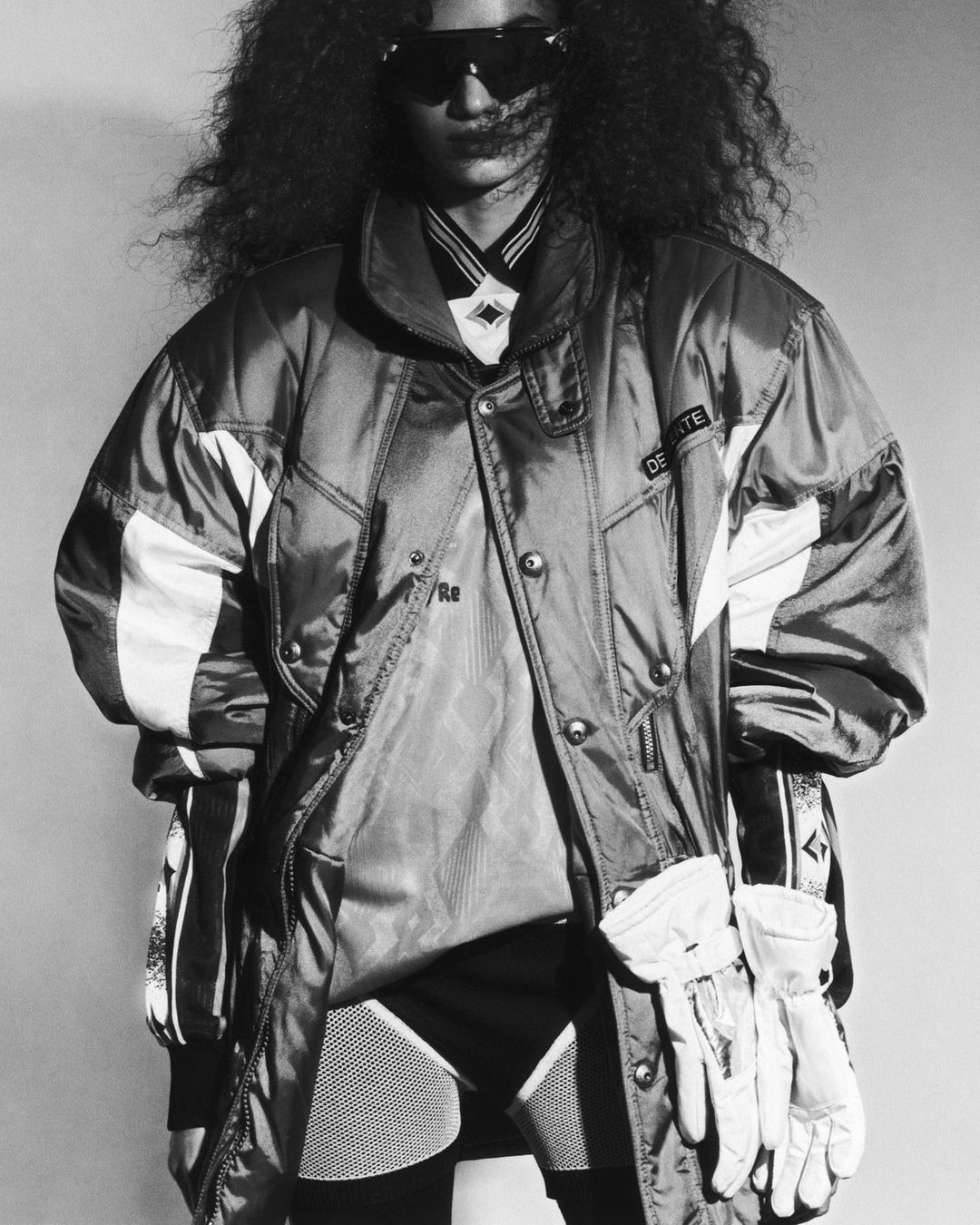 Monochrome shot of a person with curly hair wearing a puffy ski jacket and sunglasses in front of a neutral backdrop in a studio setting.