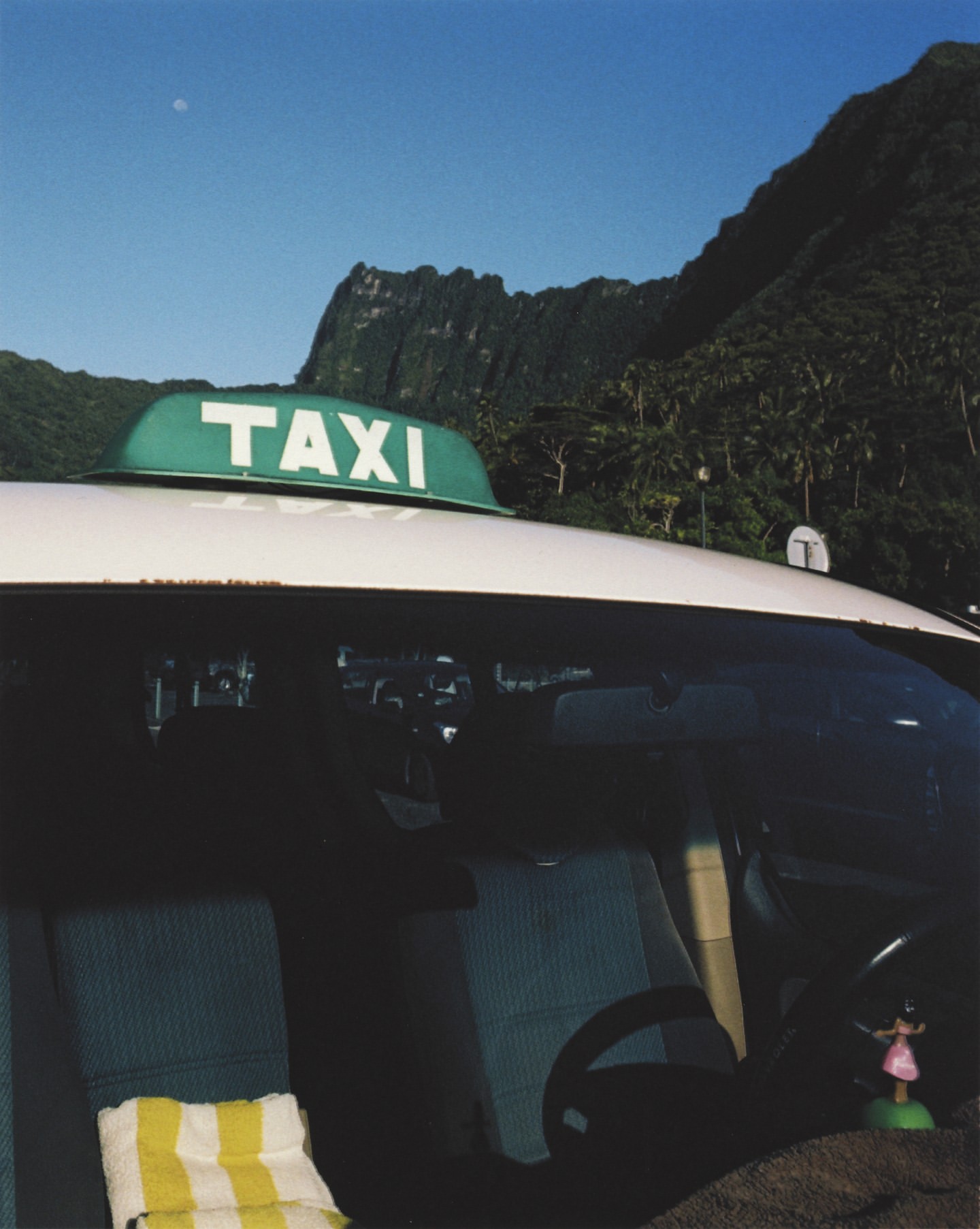 Taxi car parked outdoors in a tropical landscape featuring a view of mountains and trees under a bright blue sky in daytime.