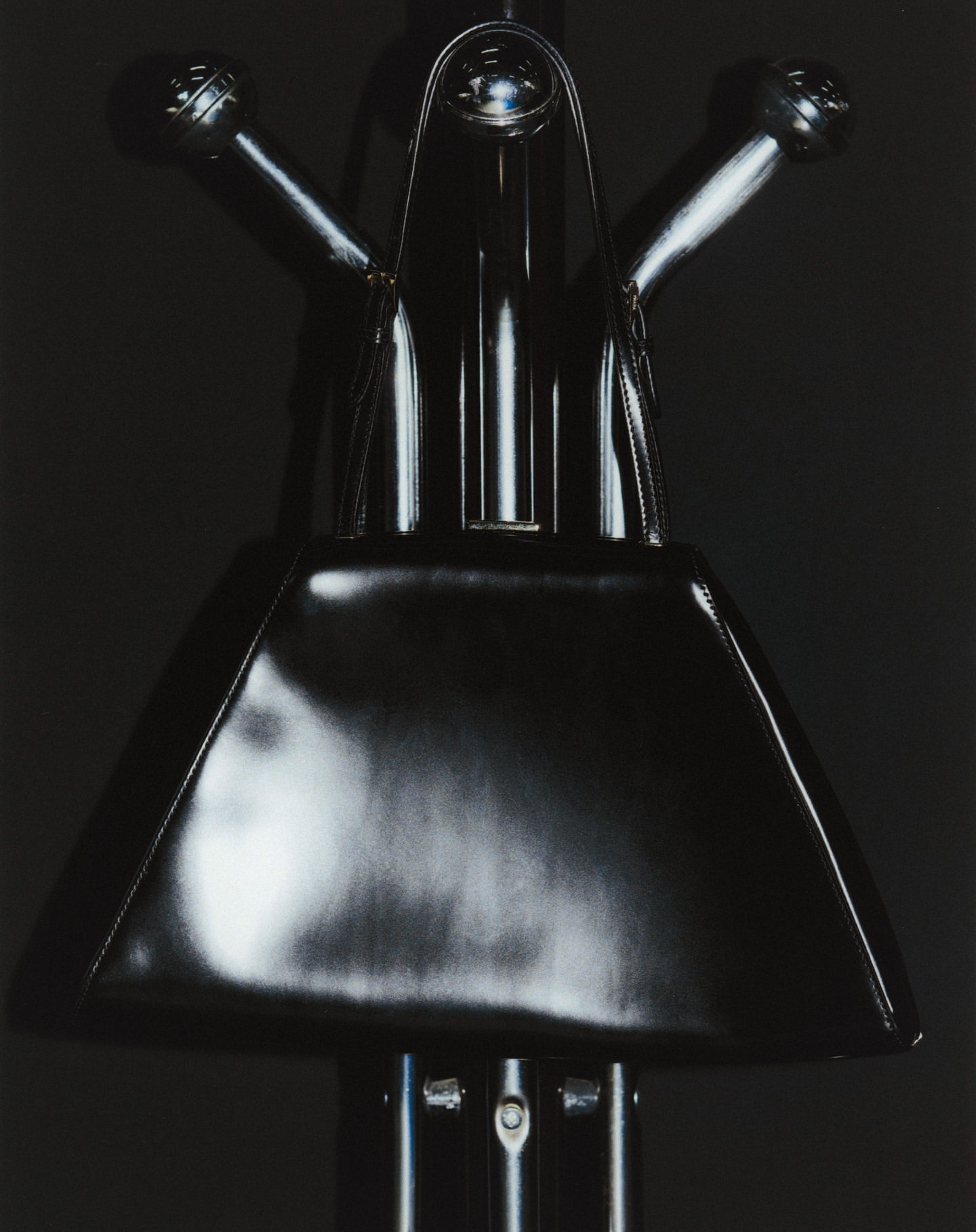 Monochrome still life of a shiny black handbag on a metallic stand against a dark background, creating a stark and sleek visual.
