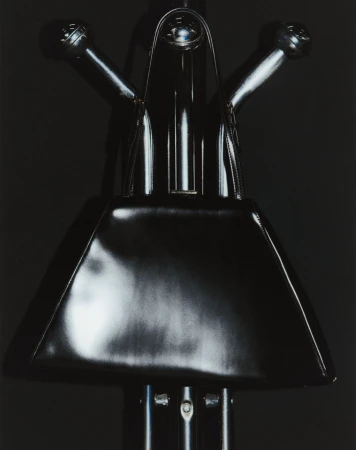 Monochrome still life of a shiny black handbag on a metallic stand against a dark background, creating a stark and sleek visual.