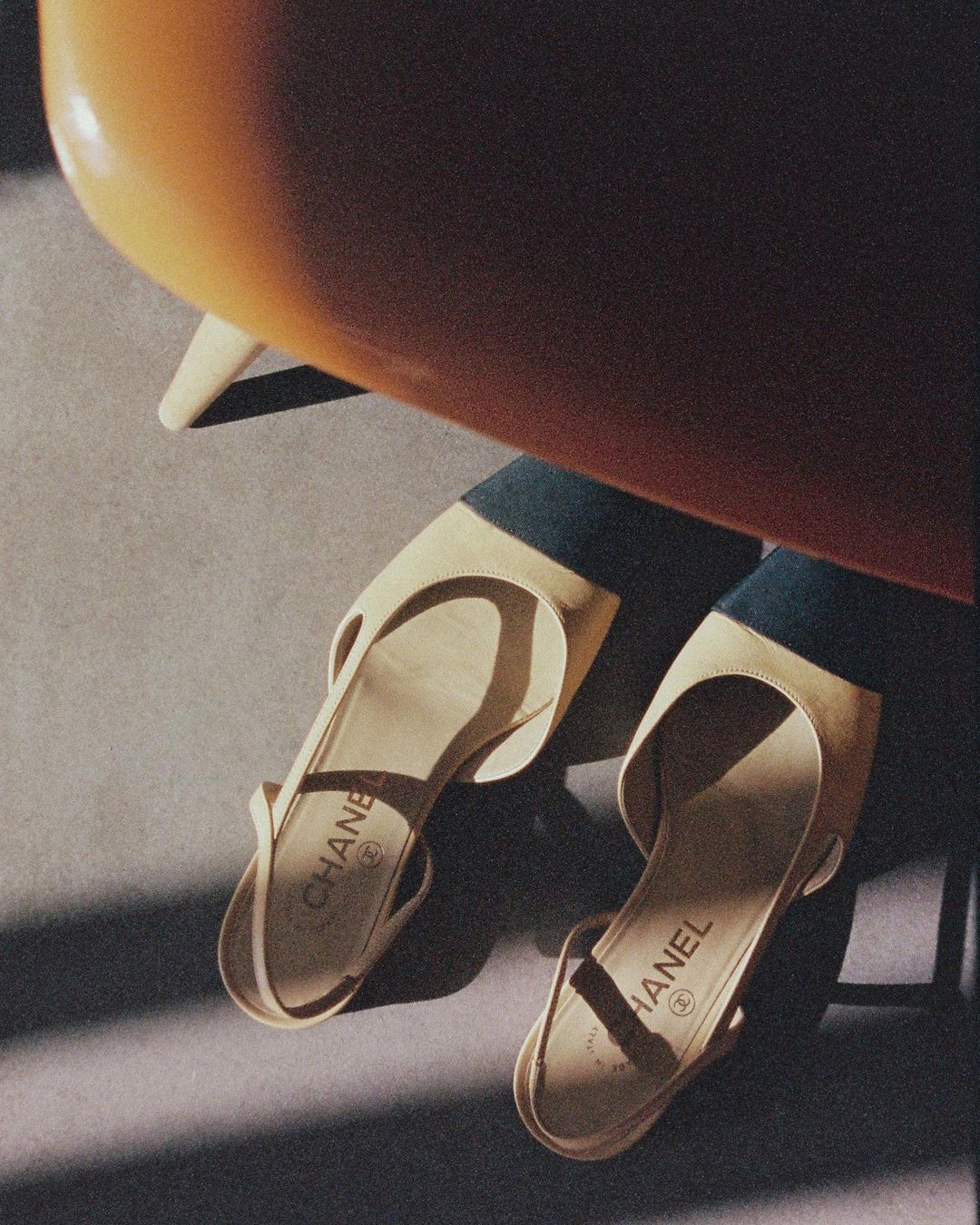 Two beige Chanel slingback shoes are angled on a grey surface, shadows cast by an orange chair above, creating a minimalist, fashionable still life.