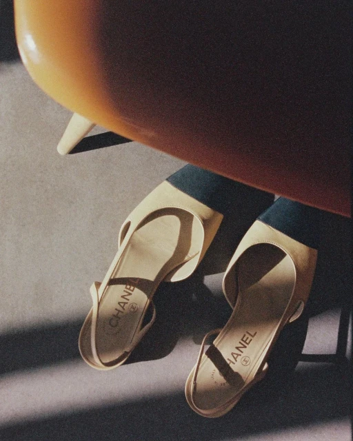 Two beige Chanel slingback shoes are angled on a grey surface, shadows cast by an orange chair above, creating a minimalist, fashionable still life.