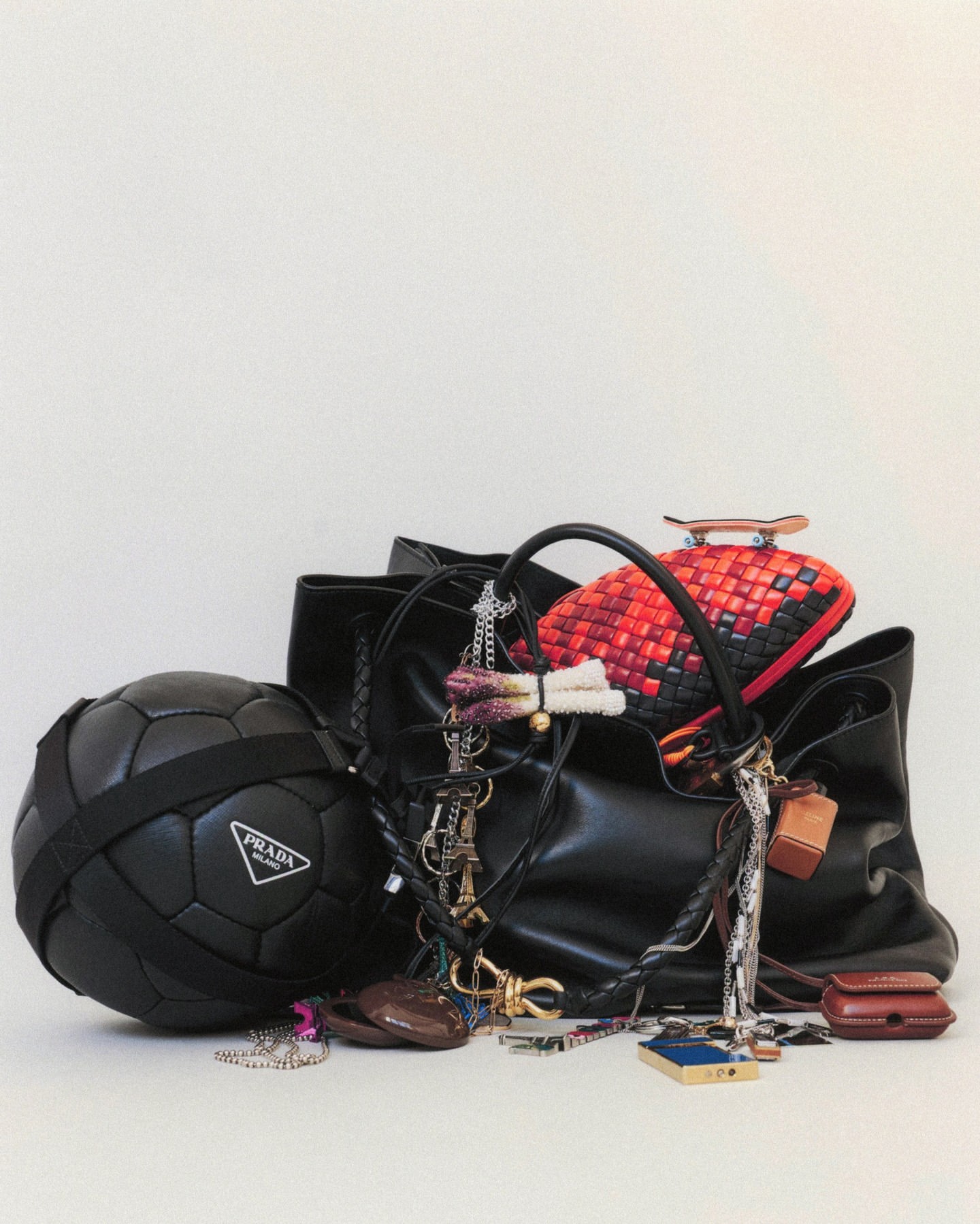 Still life composition featuring a black Prada soccer ball, an open black leather tote bag, and an assortment of accessories against a white backdrop.