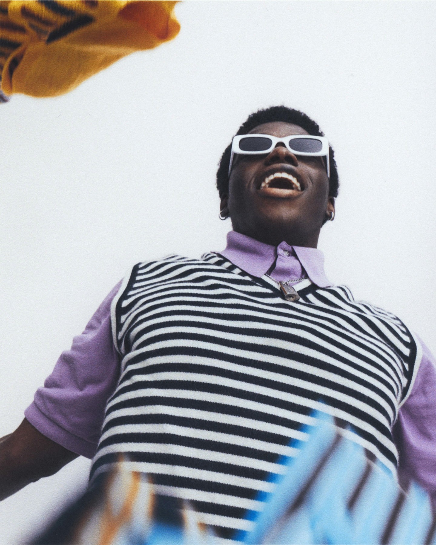 Low-angle shot of a stylish young Black man wearing sunglasses and a striped sweater vest, looking up with an open mouth in a bright indoor setting.