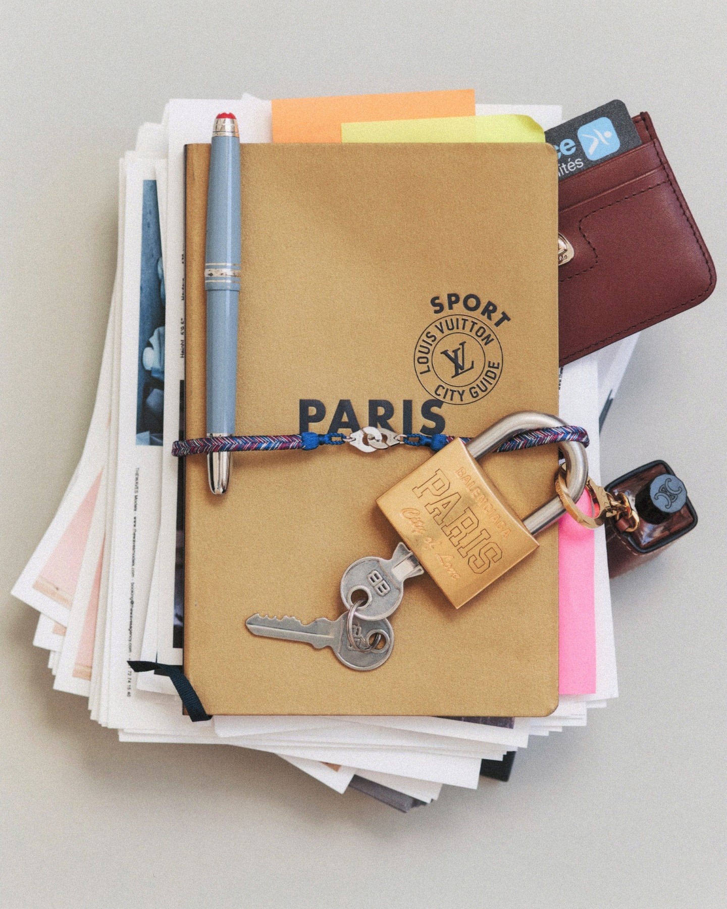 Close-up flat lay of a brown Louis Vuitton city guide notebook on a stack of papers with a silver pen, keys, and wallet.