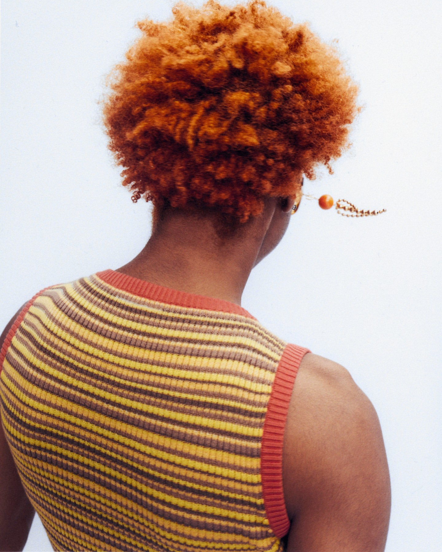 Back view of a person with short copper-colored hair, wearing a striped yellow and brown knit tank top against a bright white backdrop.