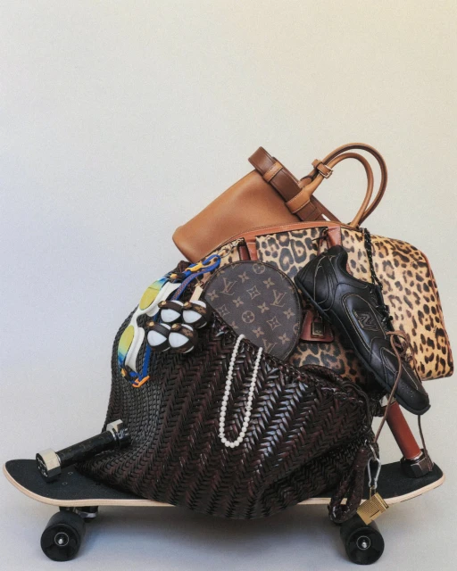 A collection of handbags and a shoe are arranged on a skateboard against a white backdrop, creating an artistic fashion still life.