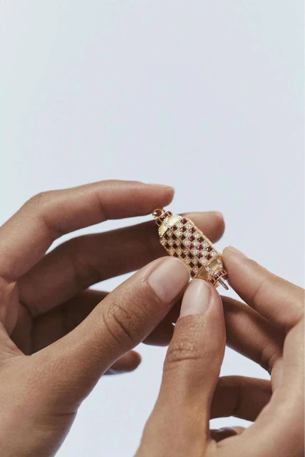 Close-up of hands holding a gold pendant featuring a checkered pattern with ruby accents, showcased against a clean, white background.