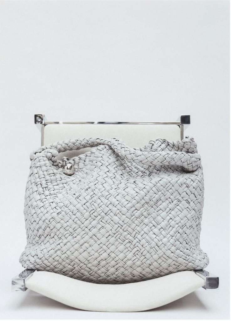 Woven leather handbag in grey and black sits atop a white cushioned chair against a white background in a fashion product studio shot.