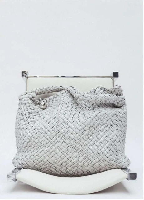 Woven leather handbag in grey and black sits atop a white cushioned chair against a white background in a fashion product studio shot.