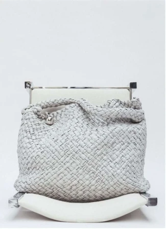 Woven leather handbag in grey and black sits atop a white cushioned chair against a white background in a fashion product studio shot.