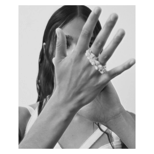 Black-and-white fashion shot of a person with pale skin and dark hair, posing with their hand up, wearing a beaded pearl ring.