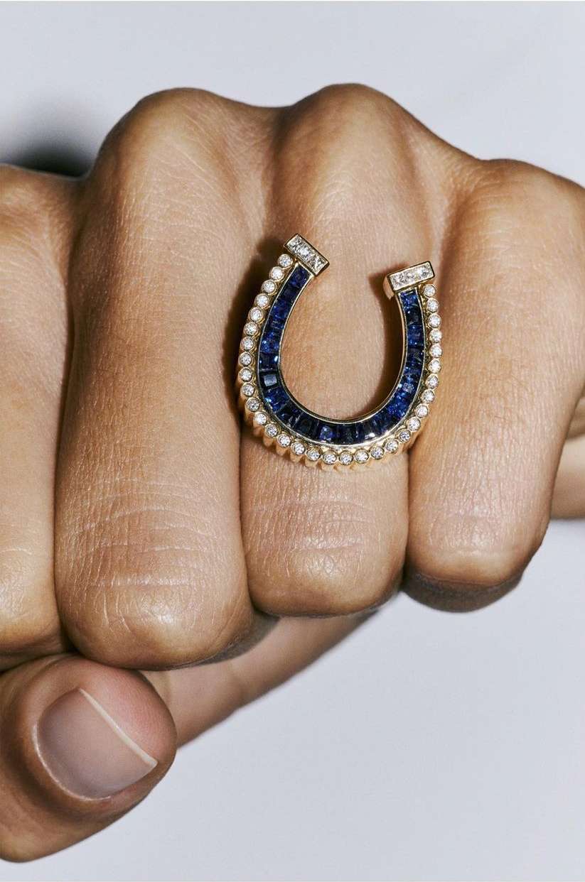 Close-up of a hand wearing a gold, blue and pearl ring in a horseshoe shape against a neutral background.