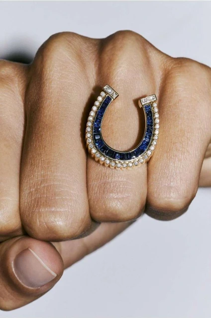 Close-up of a hand wearing a gold, blue and pearl ring in a horseshoe shape against a neutral background.