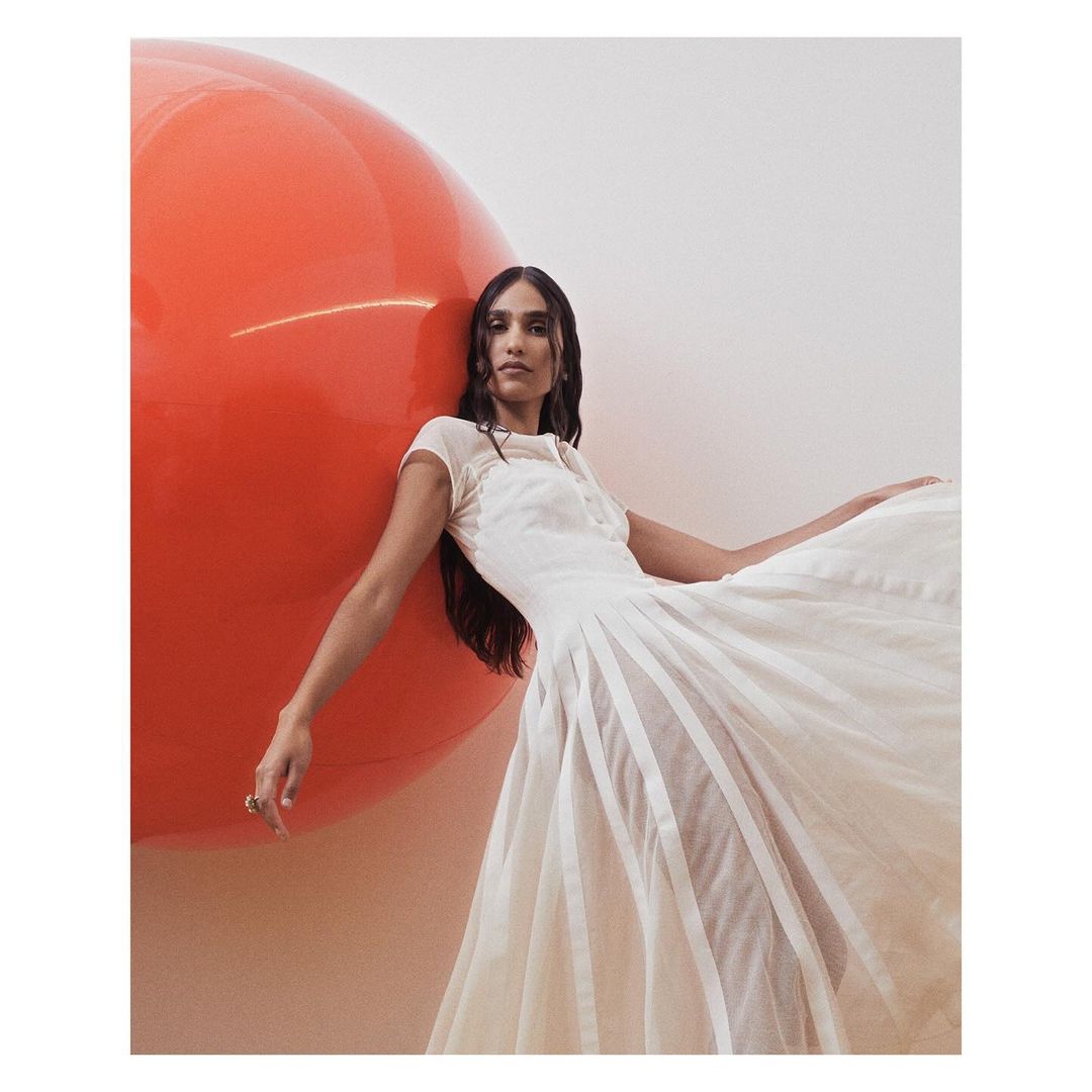 Adult woman posing in a light-colored gown next to a large, glossy orange balloon in an indoor setting. Fashion, beauty, and studio photography.
