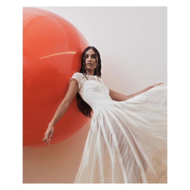 Adult woman posing in a light-colored gown next to a large, glossy orange balloon in an indoor setting. Fashion, beauty, and studio photography.