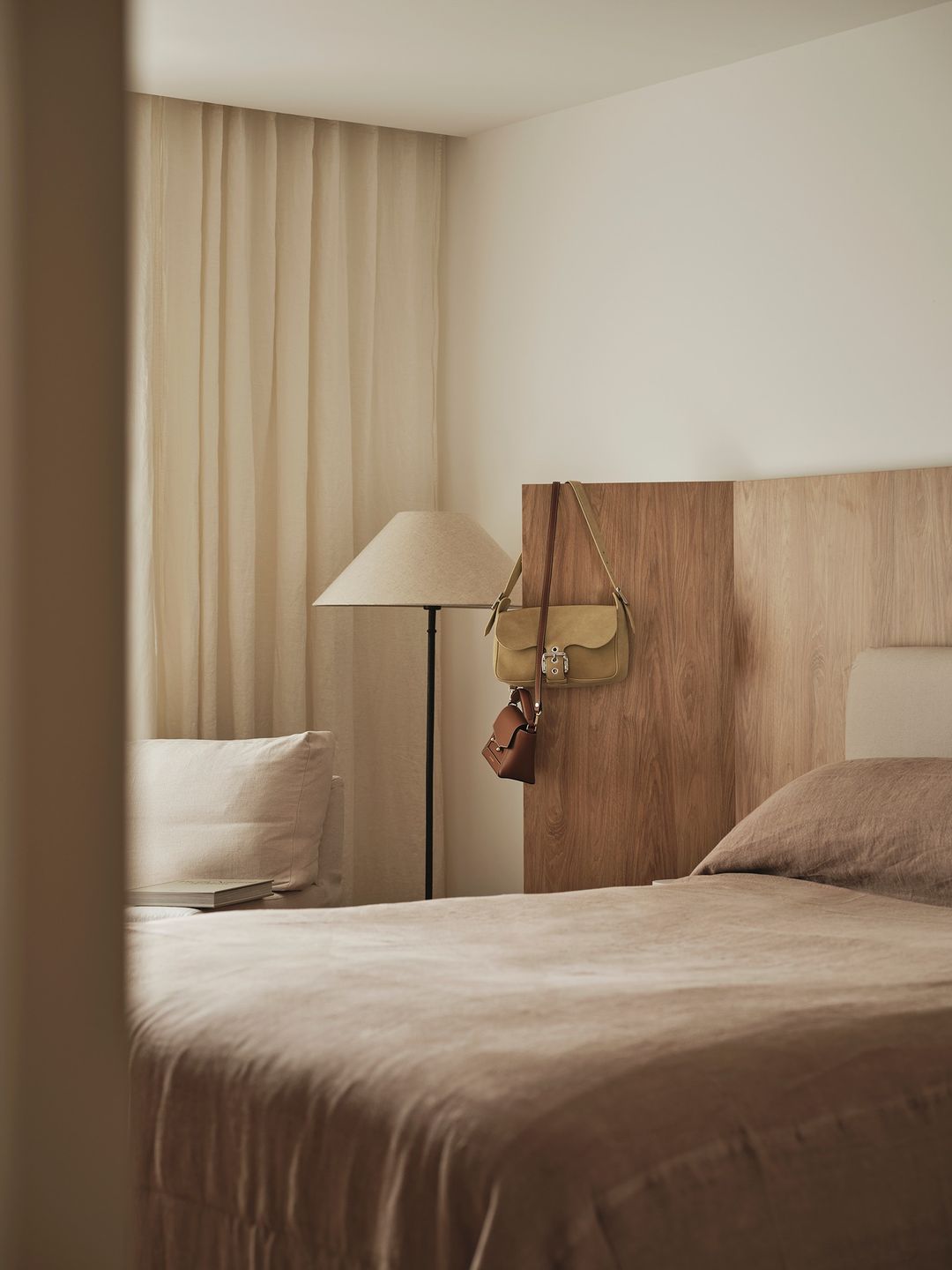 Bedroom scene with a bed covered in taupe linen, a lamp, and handbags hanging on the headboard. Cream curtains hang in the background.