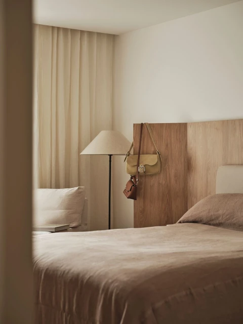 Bedroom scene with a bed covered in taupe linen, a lamp, and handbags hanging on the headboard. Cream curtains hang in the background.