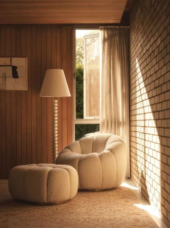 Interior of a living space with a cream-colored, rounded armchair and ottoman on a patterned rug, alongside a floor lamp near a window.