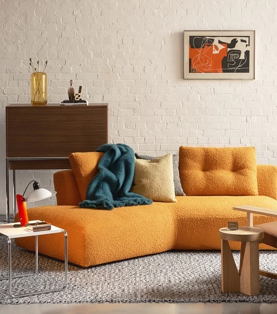 Living room interior featuring a cozy orange sofa with a teal throw blanket, complemented by modern furniture and decor against a white brick wall.