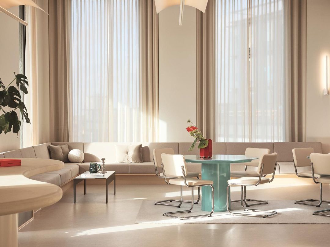 Brightly lit interior with a modern design featuring a round table surrounded by chairs, a curved sofa, and sheer curtains.
