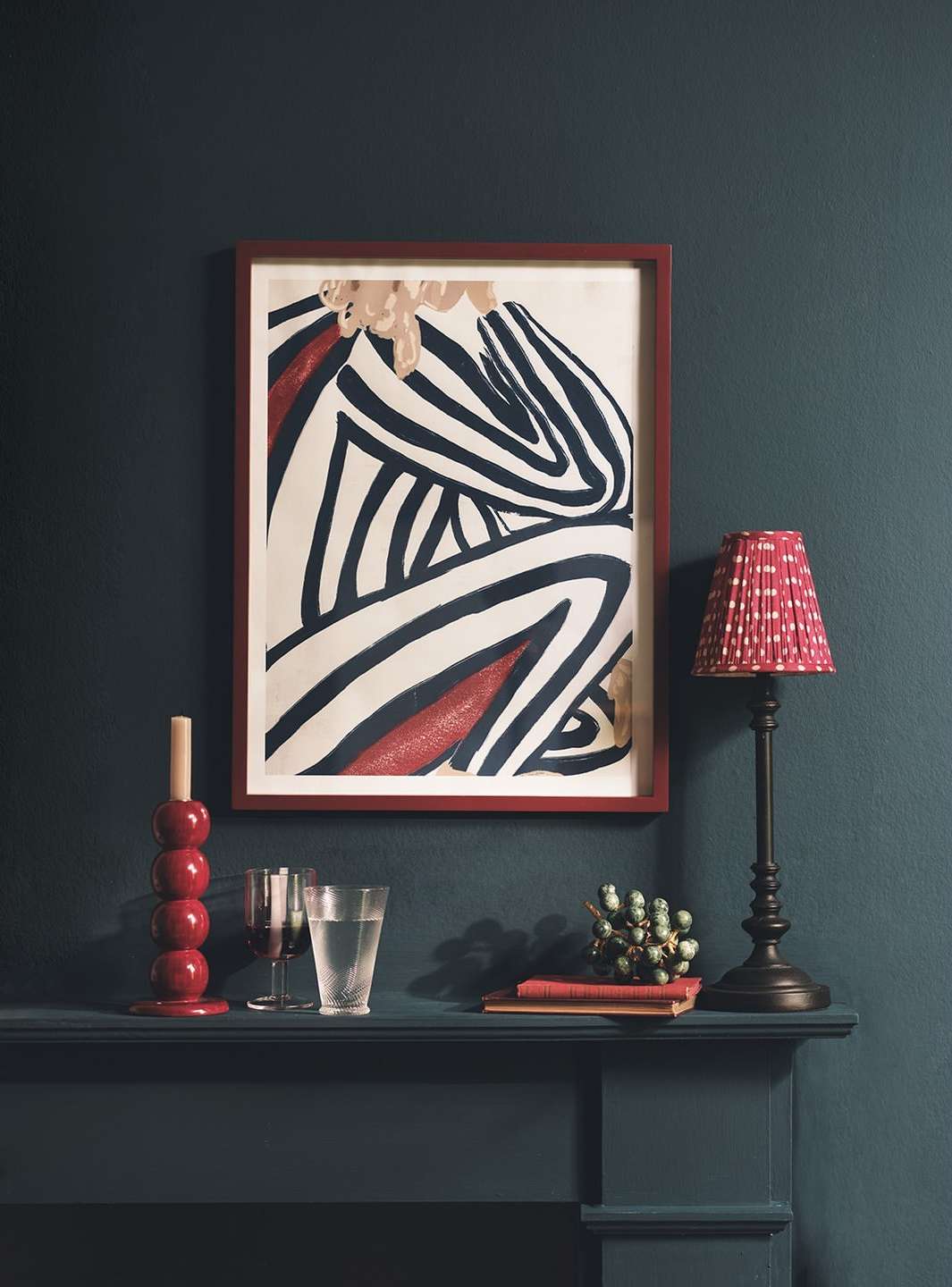 Abstract striped figure art print on wall, lamp with shade, candle holder, glass of water and books on mantle against teal wall.