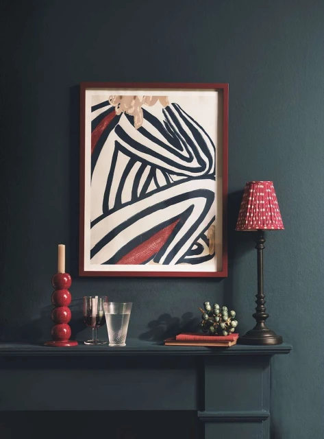 Abstract striped figure art print on wall, lamp with shade, candle holder, glass of water and books on mantle against teal wall.