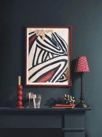 Abstract striped figure art print on wall, lamp with shade, candle holder, glass of water and books on mantle against teal wall.