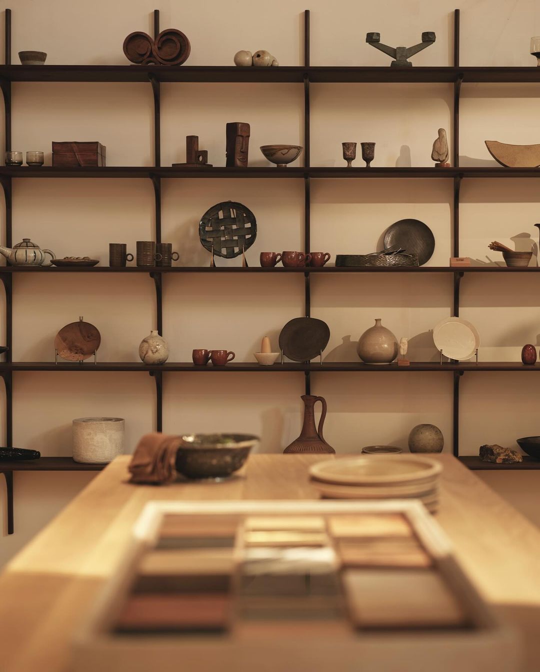 Display shelves of neutral ceramic dishes, bowls, and decorative items. Wooden table in foreground with rectangular display of samples.