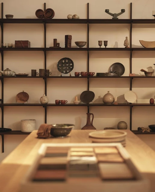 Display shelves of neutral ceramic dishes, bowls, and decorative items. Wooden table in foreground with rectangular display of samples.